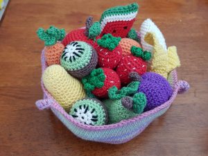 Un bol de dinette au crochet coloré rempli de divers fruits crochetés - fraises, kiwis, raisins, banane, pastèque, carotte et courge - tous disposés avec art sur une surface en bois.