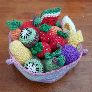 Un panier crocheté coloré rempli de fruits crochetés, notamment des fraises, des kiwis, de la pastèque, de la banane, des raisins et plus encore, présentés sur une table en bois.