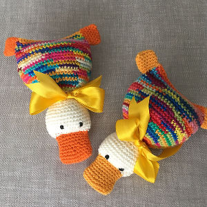 Deux canards colorés au crochet avec des becs et des pieds orange, des yeux noirs et des nœuds de ruban jaune autour du cou, posés sur un fond en tissu gris clair.