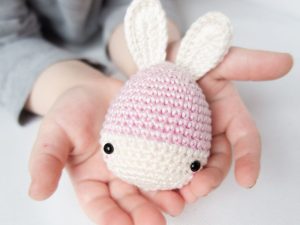 Un enfant tient un petit doudou au crochet facile : un lapin crocheté à la main avec des oreilles blanches, des yeux noirs en forme de bouton et un corps rose et blanc.