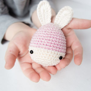 Un enfant tient un petit lapin au crochet avec des oreilles blanches, un corps rose et blanc et des yeux noirs en forme de bouton dans ses mains en coupe.