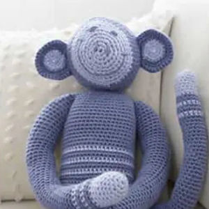 Un singe en peluche violet et bleu crocheté avec de grandes oreilles et une longue queue rayée est assis sur un coussin texturé blanc.