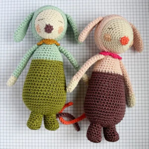Deux peluches crochetées à la main, aux oreilles tombantes et aux yeux fermés, sont posées sur une feuille de papier millimétré. L'une est verte et bleue, l'autre rose et marron, toutes deux avec un corps rond et de petits membres.