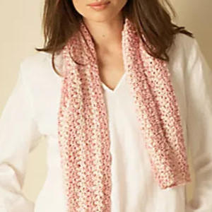 Une personne porte un haut blanc et un foulard rose clair au crochet drapé autour du cou. Le foulard présente un motif texturé, et ses cheveux bruns reposent sur ses épaules.