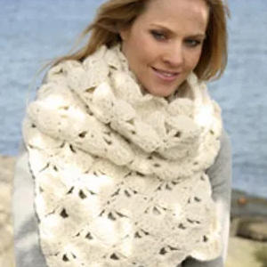 Une femme aux cheveux blonds portant une grande écharpe épaisse au crochet de couleur crème se tient à l'extérieur près d'un plan d'eau, souriant doucement.