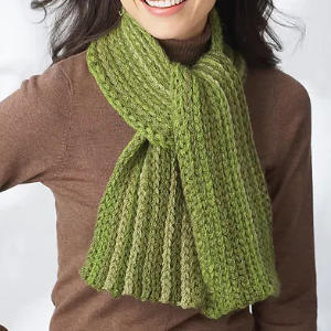 Une femme portant un pull à col roulé marron et une écharpe verte épaisse et côtelée avec un motif texturé, souriant avec ses cheveux noirs lâchés.
