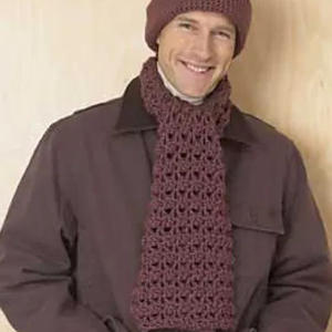 Une personne souriante portant une veste marron, un chapeau marron au crochet assorti et une grosse écharpe marron au crochet, debout sur un fond de couleur claire.