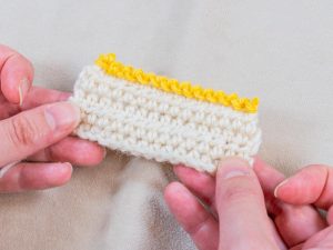 Deux mains tiennent un petit morceau de tissu crocheté avec une base crème et un bord jaune décoratif, terminé par un point d'écrevisse au crochet, présenté sur un fond beige clair.