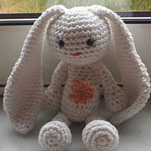 Un lapin blanc au crochet avec de longues oreilles tombantes, des yeux noirs, un petit sourire rose et une fleur couleur pêche sur sa poitrine est assis sur un rebord de fenêtre.