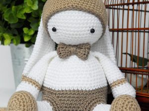 Un lapin au crochet avec de longues oreilles, des yeux en bouton noir, un nœud papillon beige et des accents beiges sur ses oreilles, ses bras et ses jambes est assis devant une plante et une cage en fil de fer marron.