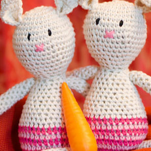 Deux poupées lapin blanches crochetées au dos rayé rose sont assises côte à côte, chacune avec un nez rose et des yeux noirs cousus. Une carotte orange est placée entre elles, sur fond rouge.