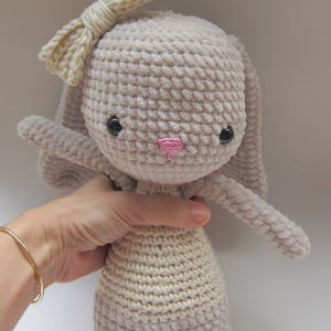 Main tenant un lapin beige clair crocheté, aux longues oreilles, au nez rose, aux yeux noirs et orné d'un nœud crème. D'apparence simple et artisanale, il est tourné vers l'avant.