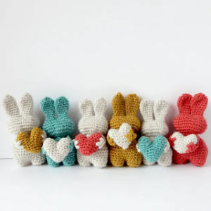 Six lapins crochetés de différentes couleurs pastel sont alignés face vers l'arrière, chacun tenant un petit cœur crocheté de différentes couleurs alors qu'ils sont assis sur un fond blanc uni.