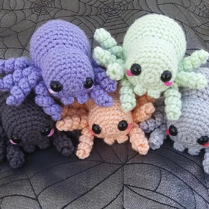 Cinq peluches d'araignées colorées au crochet avec de grands yeux noirs et des joues roses sont assises ensemble sur un fond en tissu sombre à motif de toile d'araignée.
