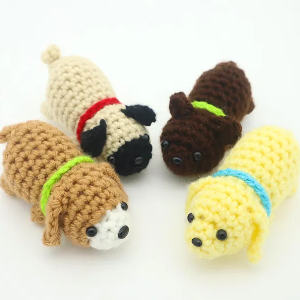 Quatre petits jouets pour chiens crochetés de différentes couleurs (beige, marron, jaune et beige), chacun avec un collier coloré, sont disposés sur un fond blanc.