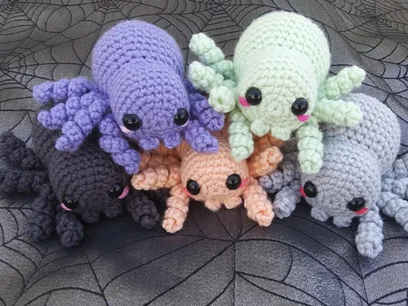 Cinq peluches araignées crochetées aux couleurs pastel, avec de grands yeux noirs et de petites joues roses, disposées sur un tissu noir à motif toile d'araignée. Parfaites pour les fans de mini amigurumi ! Un tutoriel gratuit est inclus pour créer le vôtre !.
