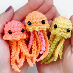Trois petites méduses colorées, crochetées au crochet et aux yeux noirs, sont délicatement tenues dans les mains. Orange, rose-orange et jaune, elles sont toutes dotées de tentacules recourbés.