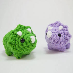 Deux petits jouets dinosaures au crochet, l'un vert et l'autre violet, chacun avec des cornes blanches et des yeux noirs, sont placés côte à côte sur un fond blanc uni.