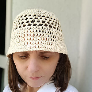 Une femme aux cheveux bruns raides regarde vers le bas tout en portant un chapeau seau au crochet de couleur crème et une chemise blanche, debout devant un mur de couleur claire.