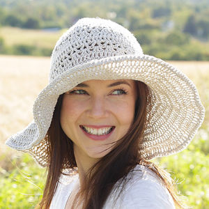 Une femme souriante aux longs cheveux bruns porte un chapeau de soleil à large bord en crochet et un haut blanc, debout à l'extérieur dans un champ herbeux ensoleillé avec des arbres en arrière-plan.