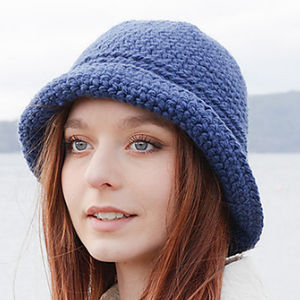 Une jeune femme aux longs cheveux auburn porte un chapeau bleu au crochet et un manteau de couleur claire, debout à l'extérieur avec un arrière-plan flou d'eau et de montagnes.