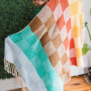 Une personne tient une couverture en tricot à carreaux, aux nuances bleu sarcelle, beige, orange et jaune. La couverture, bordée de franges, est disposée sur un fond de feuillage vert.