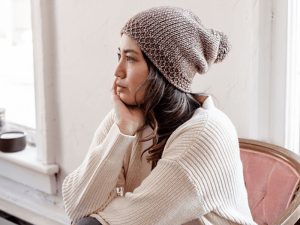 Une femme portant un bonnet en tricot et un pull couleur crème, peut-être inspiré d'un modèle gratuit de crochet tunisien, est assise sur une chaise rose, posant son menton sur sa main et regardant pensivement par la fenêtre.