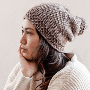 Une femme vêtue d'un pull crème et d'un bonnet tricoté est assise de profil, le visage posé sur sa main, l'air pensif.