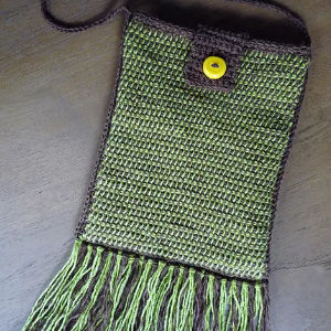 Un sac vert et marron foncé fait main au crochet avec une fermeture à bouton jaune, une longue bandoulière et une frange verte le long du bord inférieur, posé à plat sur une surface sombre.