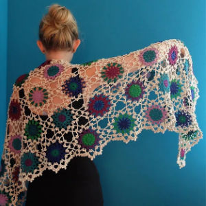 Une personne aux cheveux blonds en chignon porte une tenue noire et affiche un châle au crochet avec des motifs circulaires colorés sur un fond bleu sarcelle uni.