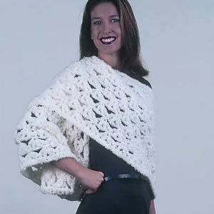 Une femme sourit en portant un poncho épais, blanc et crocheté sur une tenue noire, avec sa main droite dans sa poche, debout sur un fond clair uni.