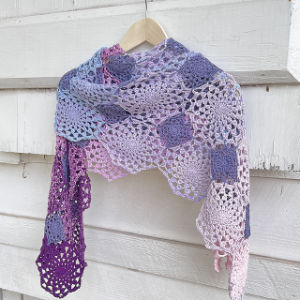 Un châle au crochet avec des motifs hexagonaux dans des tons de violet, de lavande et de rose est suspendu à un cintre en bois contre un mur en bois blanc.
