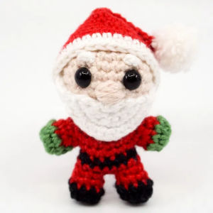 Une petite poupée de Père Noël au crochet avec un chapeau rouge, une barbe blanche, des mitaines vertes, des bottes noires et de grands yeux noirs, debout sur un fond blanc.