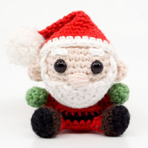 Une poupée de Père Noël au crochet avec un chapeau rouge, une barbe blanche, des yeux noirs, des mains vertes et des pieds noirs, assise sur un fond blanc.
