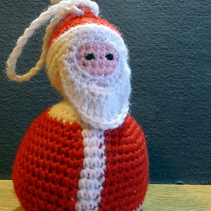 Une petite décoration de Père Noël au crochet avec un costume rouge, une bordure blanche et une barbe blanche, sur un fond sombre.