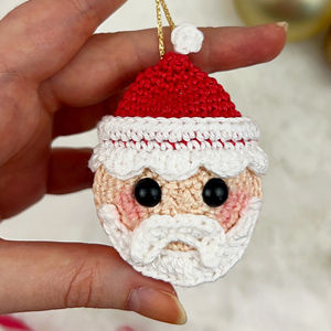 Une main tient une petite décoration de Père Noël au crochet avec un chapeau rouge, une barbe blanche et des joues roses, avec de grands yeux noirs en forme de bouton.