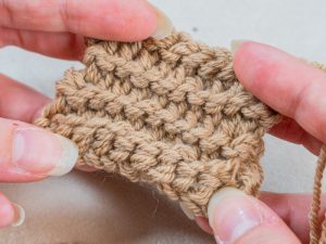 Gros plan des mains tenant un petit échantillon de fil beige texturé, présentant un motif au crochet détaillé avec des points visibles.