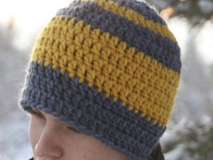 Une personne porte un bonnet épais au crochet, orné de larges rayures horizontales jaunes et grises – un look idéal inspiré d'un patron gratuit de bonnet pour homme au crochet. L'arrière-plan est flou, laissant entrevoir un paysage extérieur.