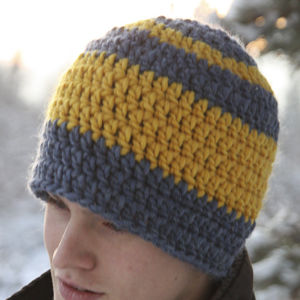 Une personne portant un bonnet crocheté à larges rayures jaunes et grises se tient dehors, dans un environnement enneigé. Seul le haut du visage est visible.