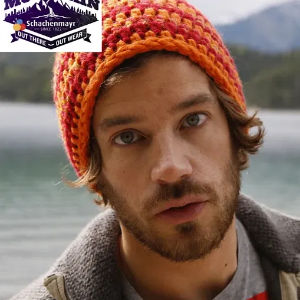 Un homme aux cheveux bruns ondulés et à la barbe coiffée d'un bonnet au crochet rouge et orange se tient dehors, près d'un lac avec des montagnes en arrière-plan. Un logo violet apparaît en haut à gauche.