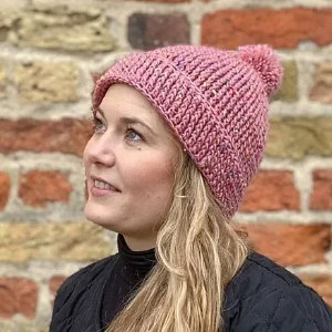 Une femme aux longs cheveux blonds porte un bonnet rose à pompon, le regard tourné vers le ciel et un léger sourire. Elle se tient devant un mur de briques.