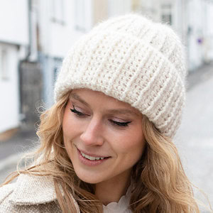Une femme aux cheveux blonds ondulés sourit tout en portant un bonnet tricoté de couleur crème et un manteau assorti, debout à l'extérieur dans une rue de la ville.