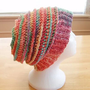 Un bonnet souple coloré, crocheté à la main, avec des motifs rayés, est présenté sur une tête de mannequin blanche, posée sur une surface en bois clair.