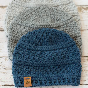 Trois bonnets crochetés sont empilés sur une surface en bois ; deux sont gris clair et un bleu foncé avec une étiquette marron. Les bonnets présentent un design texturé et à motifs.