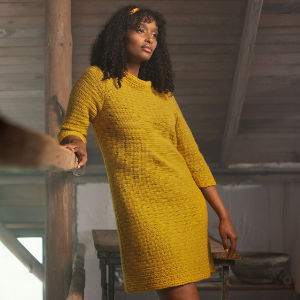 Une femme aux cheveux bouclés porte une robe jaune texturée à manches trois-quarts, debout à l'intérieur dans un décor rustique en bois, appuyée contre une poutre en bois.