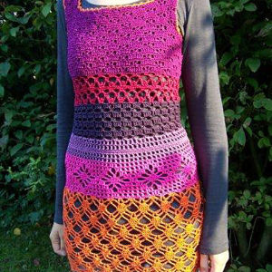 Une personne portant une robe colorée au crochet faite à la main avec des motifs violets, roses, rouges et orange sur une chemise grise à manches longues, debout à l'extérieur avec de la verdure en arrière-plan.