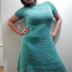 Une femme pose avec une main sur la hanche, vêtue d'une robe bleu sarcelle clair, à manches courtes, crochetée à la main avec des détails en dentelle à l'ourlet.