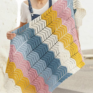 Une personne portant une chemise blanche et une salopette tient une couverture au crochet avec un motif à chevrons dans les couleurs rose, bleu, jaune et crème.