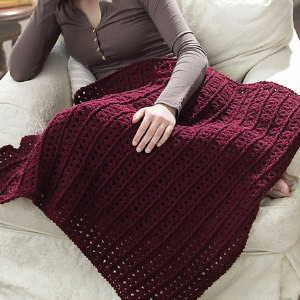 Une personne portant une chemise marron à manches longues est assise sur un canapé blanc avec une couverture texturée au crochet rouge foncé drapée sur ses genoux.