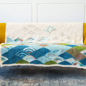Une couverture en tricot colorée avec des motifs de vagues et d'éventails en bleu, vert et crème drapée sur un canapé jaune contre un mur lambrissé blanc.